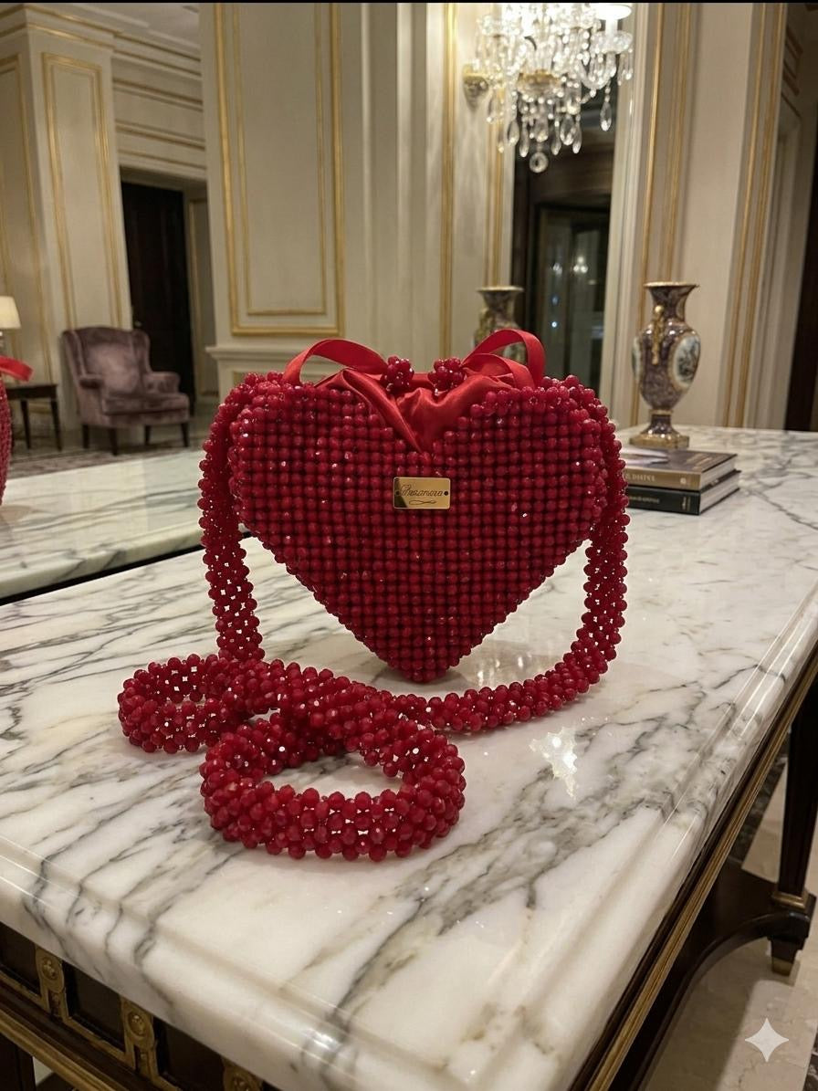 Bassmera Valentina Crimson red crystal beaded heart bag standing upright on marble table in luxury hotel setting, beaded crossbody strap laid out, red satin bow at top, gold Bassmera plaque
 